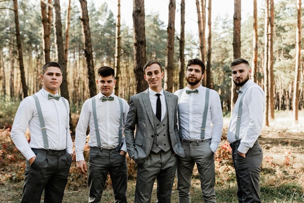 Groom with friends hold hands in pockets and look at the camera, middle-shot of a handsome groom, wedding photo session in nature, wedding in rustic style