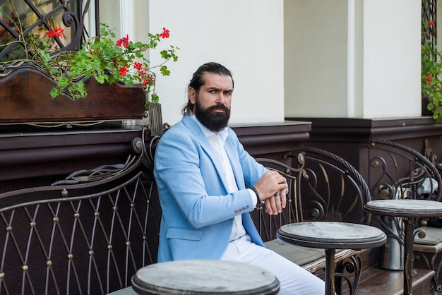 Groomed brutal hipster in stylish clothes male beauty and fashion confident businessman with beard and moustache mature man in formal elegant suit relax in cafe bride groom outdoor