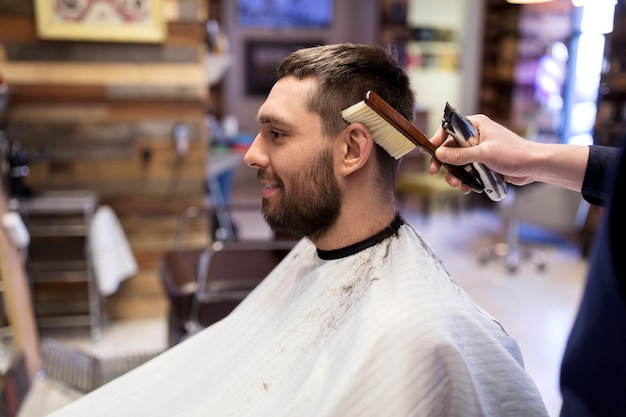 grooming, hairdressing and people concept - man and barber with trimmer and brush cleaning hair at barbershop