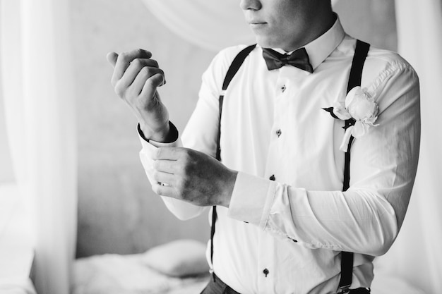 Grooms morning preparation handsome groom in bow tie suspenders getting dressed and preparing for the wedding at hotel the bridegroom wears cufflinks in a white shirt black and white portrait