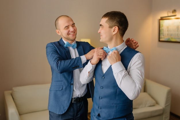 Groomsmen in vestito blu che aiuta sposo felice che si prepara al mattino per cerimonia di nozze. uomo di lusso in tuta in camera. giorno del matrimonio.