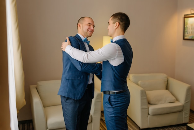 Groomsmen in blue suit helping happy groom getting ready in the morning for wedding ceremony. luxury man in suit in room. wedding day.