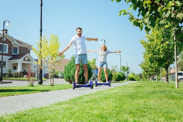 Groot plezier. Opgewonden jonge man en vrouw rijden hoverboards over het trottoir en spreiden hun handen wijd uit, genietend van de rit