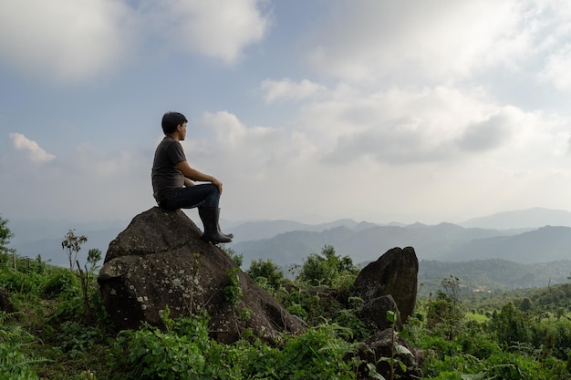 Groothoekopname van een Aziatische man die op de rots zit en kijkt naar een verre avontuurlijk alleenstaande natuur