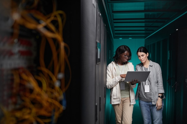 Groothoekportret van twee jonge vrouwen die een laptop gebruiken in de serverruimte tijdens het opzetten van een supercomputernetwerk, kopieer ruimte