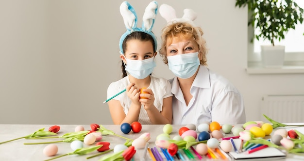 grootmoeder en kleindochter maken thuis paasdecoratie en dragen een gezichtsmasker om te beschermen tegen het coronavirus.