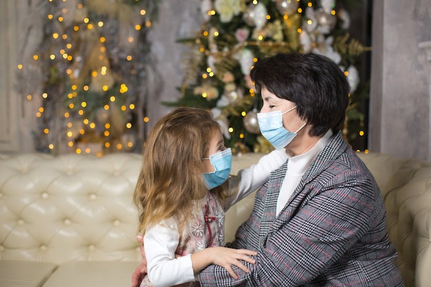 Grootmoeder en kleindochter op de bank in de woonkamer met kerstdecor knuffelen in medische maskers op hun gezichten. Een gezinsvakantie tijdens het uitbreken van coronavirus en ziekte. Nieuwjaar