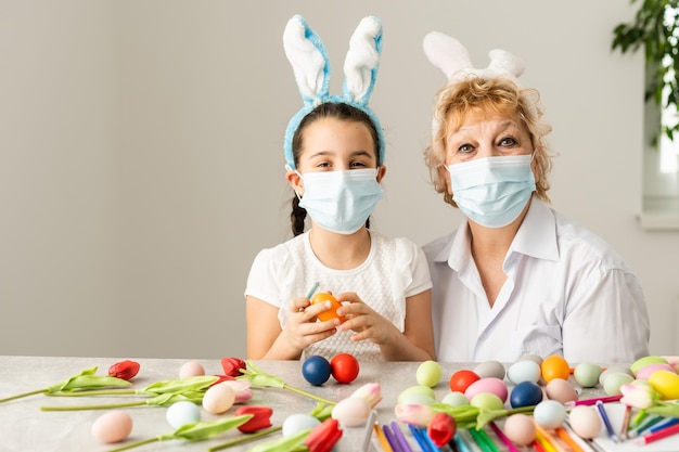 grootmoeder en kleindochter schilderen paaseieren in de keuken met kwasten, telefoon, tulpen en konijnen thuis in quarantaine met medisch masker. Coronavirus, ziekte, infectie, quarantaine, griep, operatie