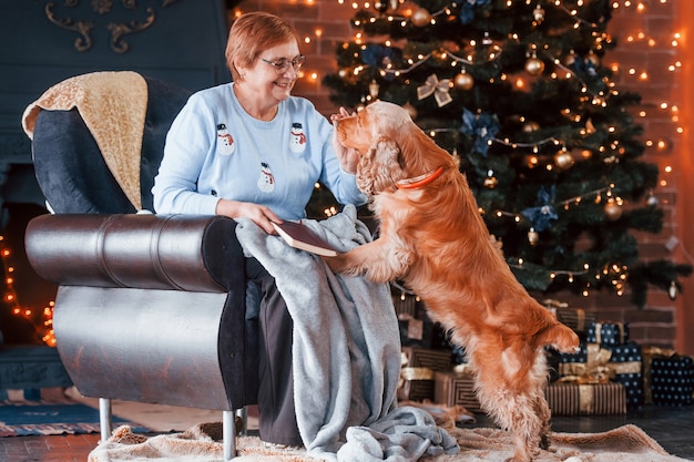 Grootmoeder met boek in de hand binnenshuis met hond in kerst ingerichte kamer.