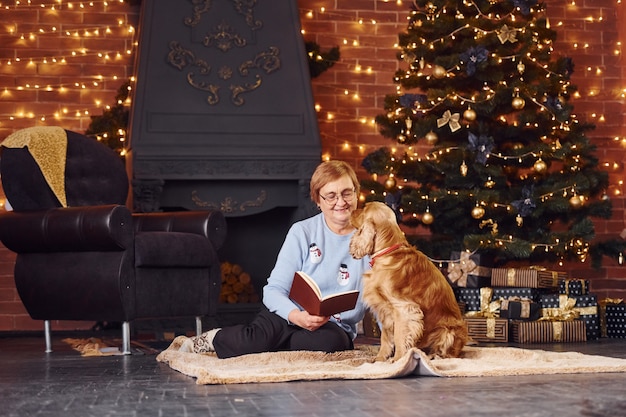 Grootmoeder met boek in de hand binnenshuis met hond in kerst ingerichte kamer.