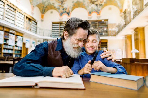 Grootvader en kleinzoon die en terwijl het zitten in oude uitstekende bibliotheek glimlachen koesteren en grappig opwindend boek lezen. opa leest een boek voor zijn kleinzoon in de bibliotheek.