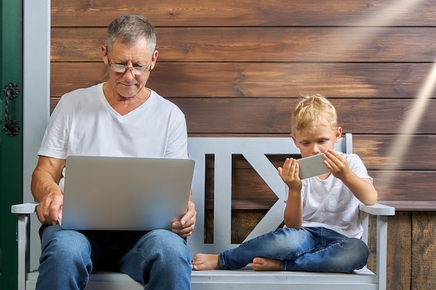 Grootvader met een laptop en een kleinzoon met een smartphone zitten op een bankje in de buurt van een houten huis Concept van het gebruik van moderne gadgets door verschillende generaties