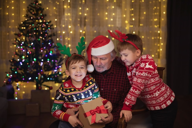 Grootvader met kerstmuts en kleinzonen openen kerstcadeau, poseren in feestelijke kamer met kerstversiering en nieuwjaarsboom. Kleine jongen is blij met zijn huidige glimlach en kijkt naar de camera.