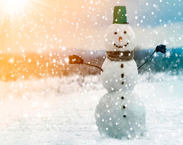 Grote glimlachende sneeuwman met emmerhoed sjaal en handschoenen op wit besneeuwd veld winter landschap vallen sneeuwvlokken en blauwe hemel kopieer ruimte achtergrond Vrolijk kerstfeest en Gelukkig Nieuwjaar groeten kaart