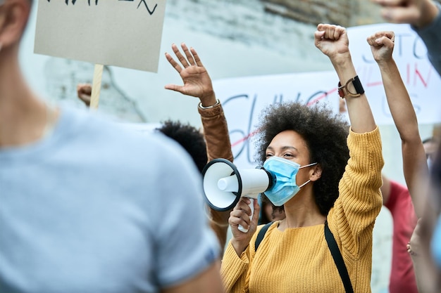 Grote groep demonstranten met opgeheven vuisten die deelnemen aan demonstraties tijdens COVID19-epidemie Focus ligt op zwarte vrouw met beschermend gezichtsmasker die door megafoon schreeuwt