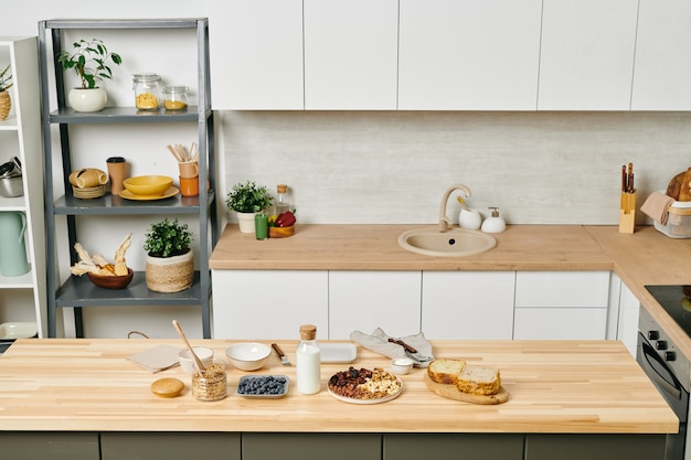 Grote moderne keuken met keukengerei op planken, witte kasten aan muren, eten en melk op houten tafel