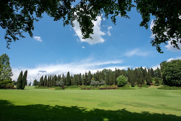 Grote open tuin met groenblijvende cipressen bekeken over een keurig onderhouden gazon van groen gras onder een zonnige blauwe hemel met wolken in een schilderachtig landschap