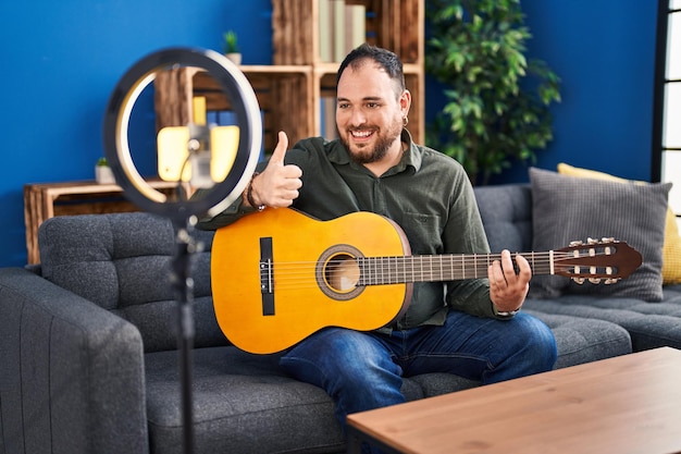 Grote Spaanse man met baard die klassieke gitaar speelt in de muziekstudio en zichzelf opneemt glimlachend gelukkig en positief, duim omhoog doet uitstekend en goedkeuringsteken