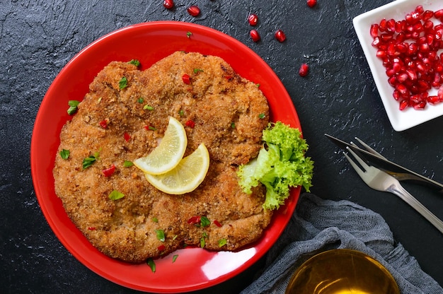 Grote Weense schnitzel op een rode plaat met citroen op een zwarte achtergrond. Vleesschotel. Bovenaanzicht, plat gelegd.