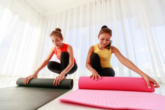 Ground level of Asian girlfriends rolling mats and looking at each other at end of yoga session in light studio