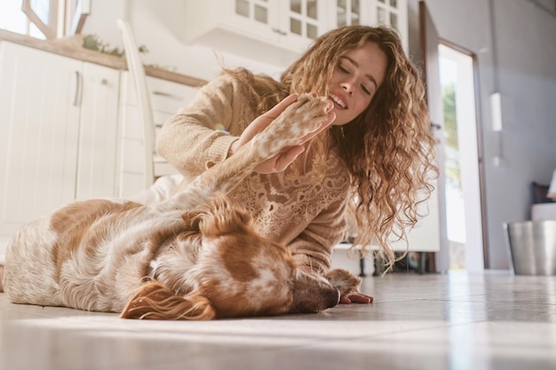 Ground level of smiling young woman with long curly hair giving high five to cute dog while playing together on kitchen floor