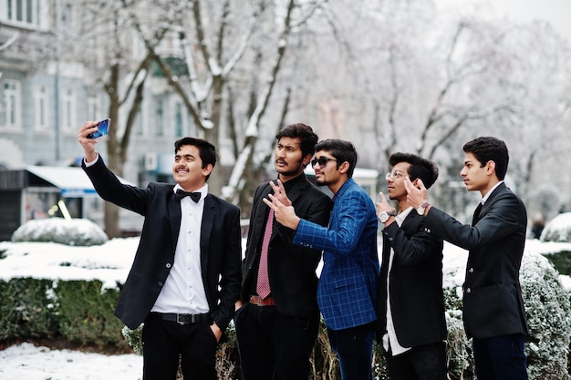 Group of 5 indian students in suits posed outdoor in winter day and making selfie on phone