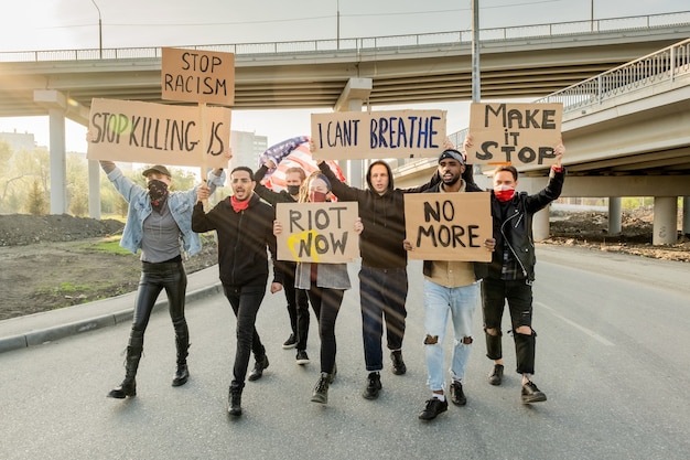 Группа разгневанных молодых многонациональных людей поднимает картонные плакаты, протестуя против расизма на уличной демонстрации