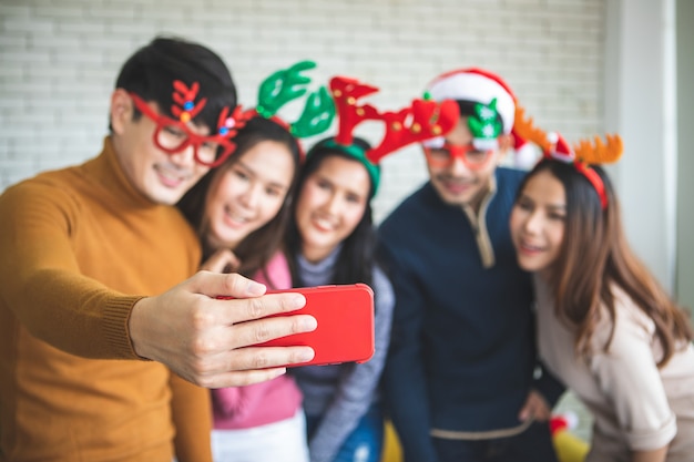 Group of asian friends taking selfie with friend together by smartphone at home during chrismas eve party or New year celebrate party. Happy winter Xmas and Happy new year party concept