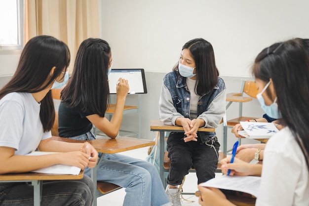 グループのアジアの大学生が保護マスクを着用して教室でプロジェクトについて話し合う