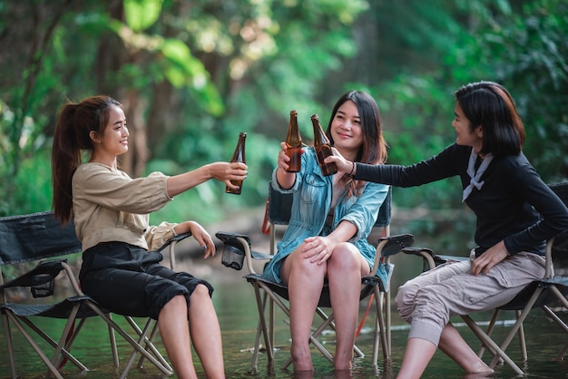 Group beautiful Asian women friends travelers relaxing in camp chairs in stream They are cheering and drinking beer during camping talking with fun and happy together