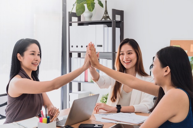 Group of beautiful  happy Asian women meeting and giving high five hand touch together in office space to discussion or brainstorm business startup project..