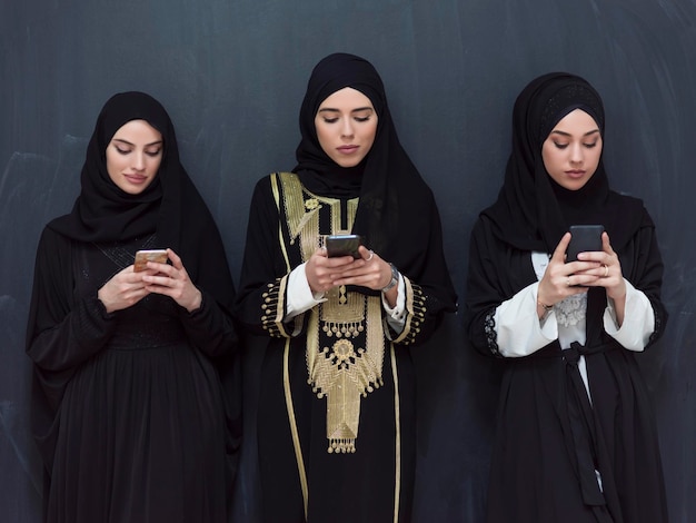 group of beautiful Muslim women in a fashionable dress with hijab using mobile phones in front of a black chalkboard representing modern Islam fashion technology and Ramadan Kareem concept. High-quali