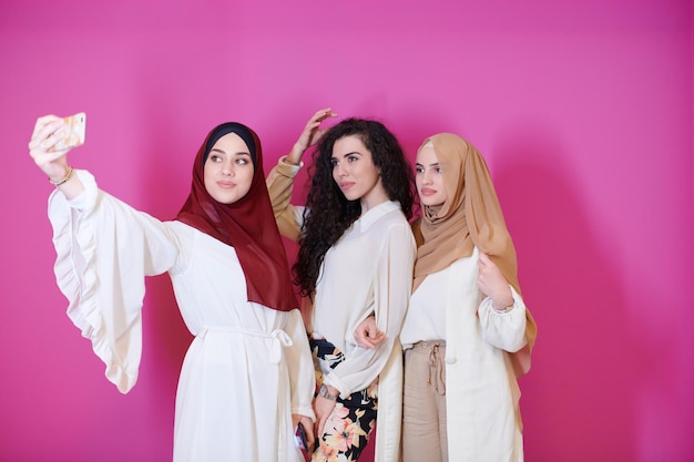 現代のイスラムファッション技術を表すピンクの背景に隔離された自撮り写真を撮りながら、携帯電話を使用してヒジャーブを着たファッショナブルなドレスを着た美しいイスラム教徒の女性2人のグループと