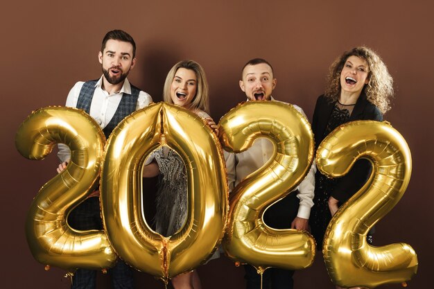 Group of beautiful well dressed party people celebrating arrival of 2022 New Year with a golden balloons on brown background