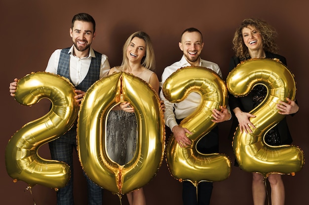 Group of beautiful well dressed party people celebrating arrival of 2022 New Year with a golden balloons on brown background