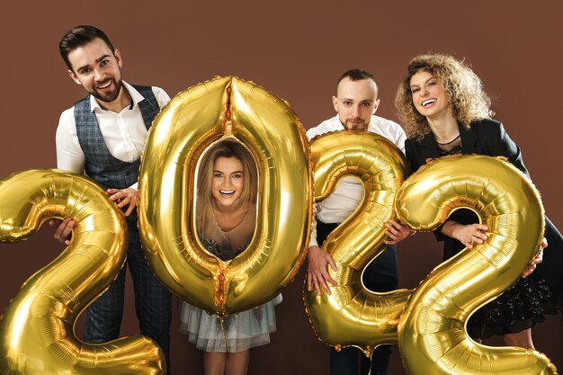 Group of beautiful well dressed party people celebrating arrival of 2022 New Year with a golden balloons on brown background