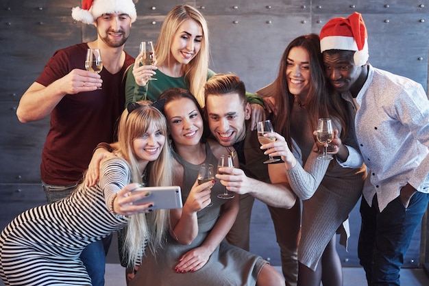 Raggruppi i bei giovani che fanno il selfie nel partito del nuovo anno, le ragazze e i ragazzi dei migliori amici che si divertono insieme, posando il concetto della gente di stile di vita. cappelli di babbo natale e bicchieri di champagne nelle loro mani