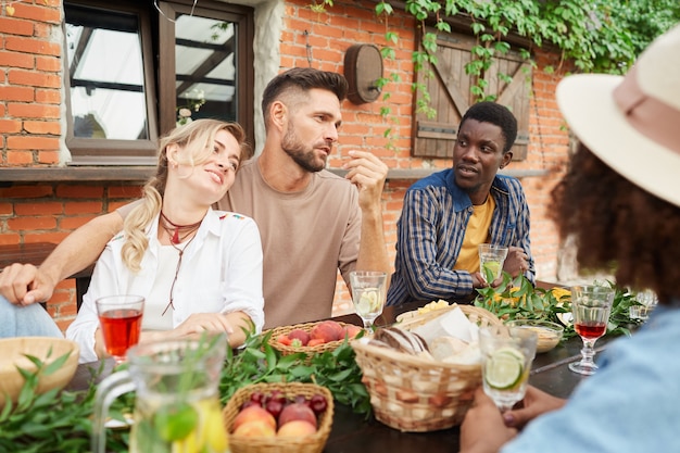 Group of beautiful young people enjoying dinner outdoors while sitting at wooden table by cottage