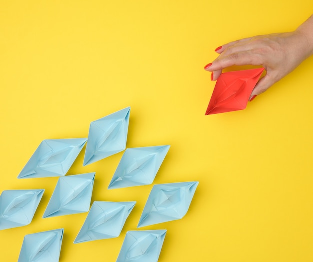 a group of blue paper boats follows a red boat in front of a yellow background