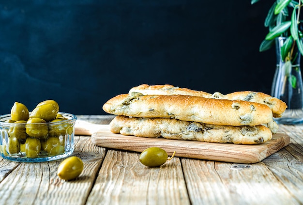 Group of bread sticks stuffed with green olives ion cutting board olive tree branch olives in bowl on wooden table with dark background