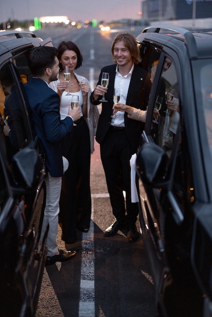 Photo group of business partners have fun together drinking alcohol while celebrating between cars on parking lot at night