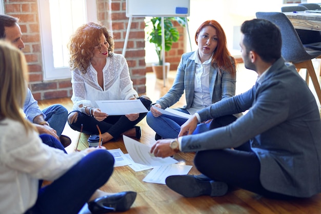 Group of business workers smiling happy and confident sitting on the floor relaxed working together speaking and reading documents at the office
