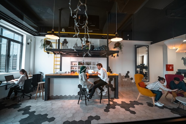 캐주얼한 다민족 기업인 그룹은 현대적인 개방형 계획 시작 사무실의 휴식 공간에서 자유 시간을 즐기면서 다양한 일을 하며 휴식을 취하고 있습니다. 고품질 사진