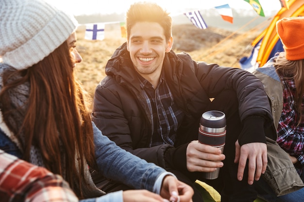 Group of cheerful young friends camping outdoors, sitting with cups and thermos