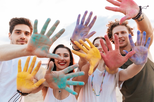 Gruppo di giovani amici allegri che si divertono in spiaggia con vernice colorata holi, mostrando le mani