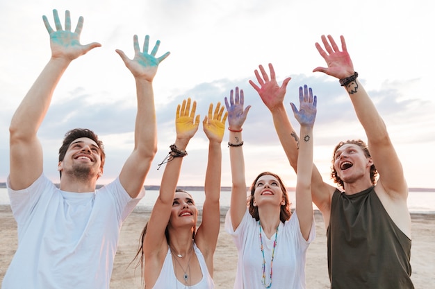 Gruppo di giovani amici allegri che si divertono in spiaggia con vernice colorata holi, mostrando le mani