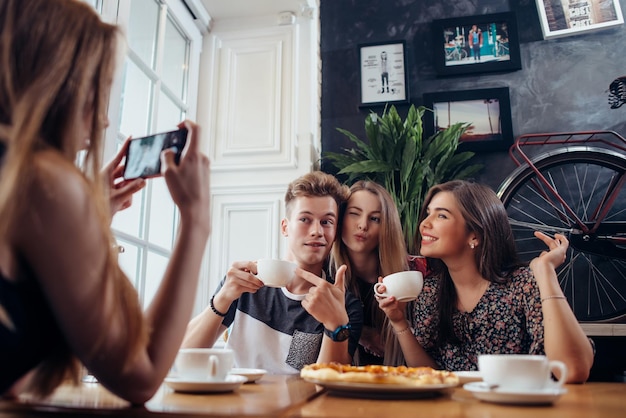 Group of cheerful young people posing in a cafe drinking coffee making faces while their friend photographing them with mobile phone