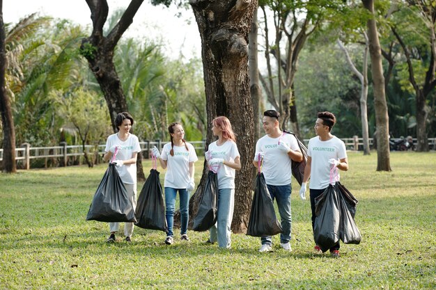 Photo group of cheerful young volunteers carrying bags of trash they picked on campus or in city park