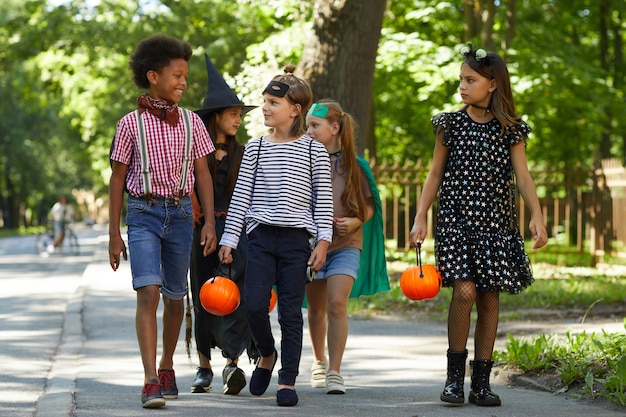 Group of children in costumes with toy pumpkins walking along the street to celebrate Halloween holiday