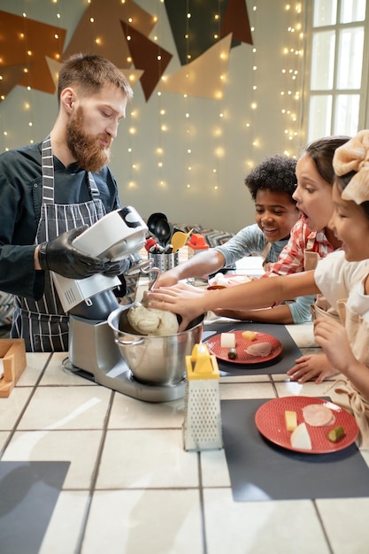 料理人の助けを借りて料理を学ぶ子供たちのグループ、彼らは台所での準備にミキサーを使用しています